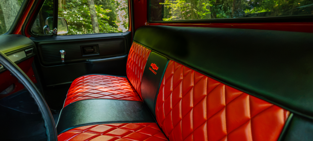A close-up view of a custom red and black quilted leather truck bench seat inside a vehicle, with a forest visible through the window.